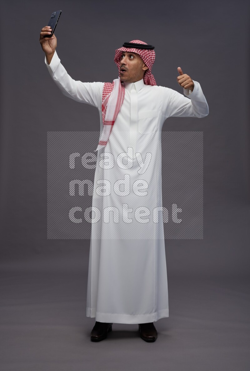 Saudi man wearing thob and shomag standing taking selfie on gray background