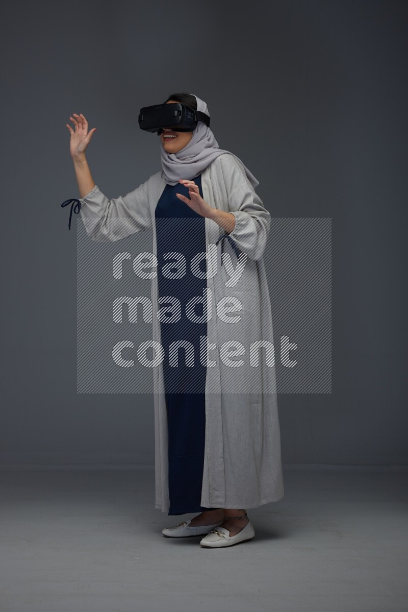 A Saudi woman wearing a light gray Abaya and head scarf standing and wearing VR eye level on a grey background