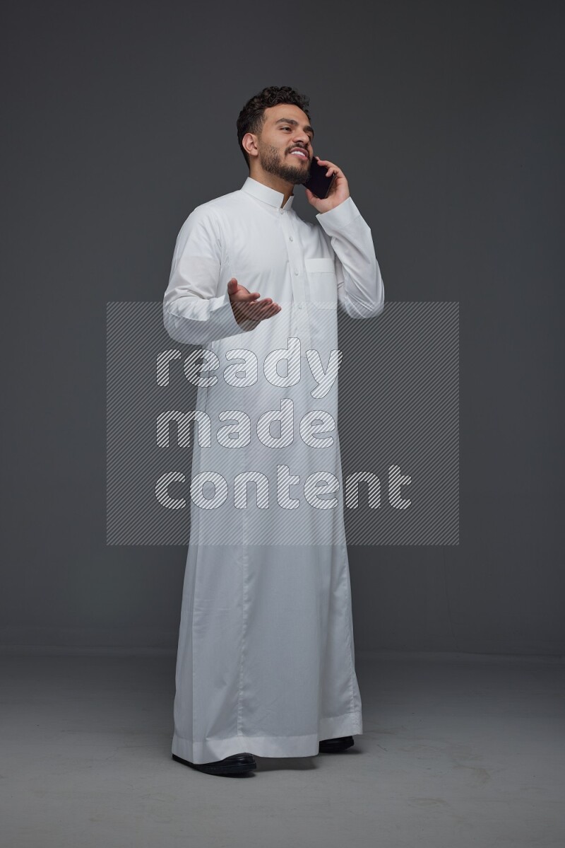 A Saudi man wearing Thobe and talking in the phone while standing and making different poses eye level on a gray background