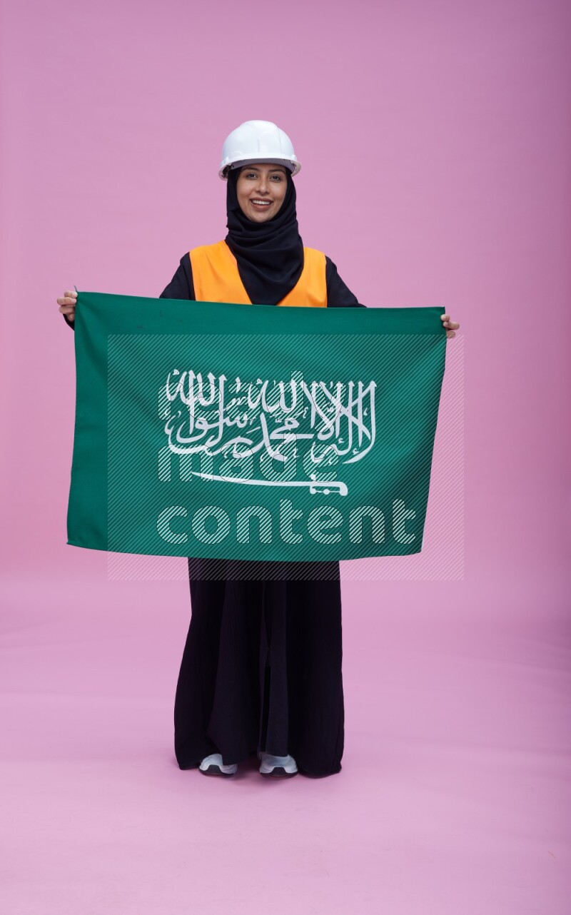 Saudi woman wearing Abaya with engineer vest and helmet standing holding Saudi flag on pink background