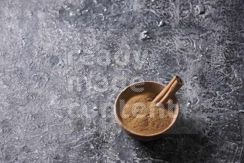Wooden bowl full of cinnamon powder and a cinnamon stick on a textured black background