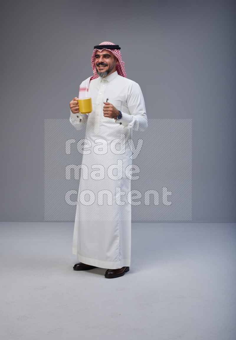 Saudi man Wearing Thob and red Shomag standing holding mug on Gray background
