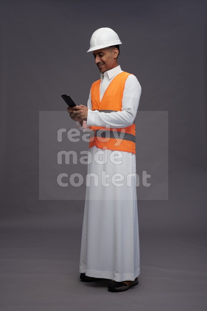 Saudi man wearing thob with engineer vest standing texting on phone on gray background