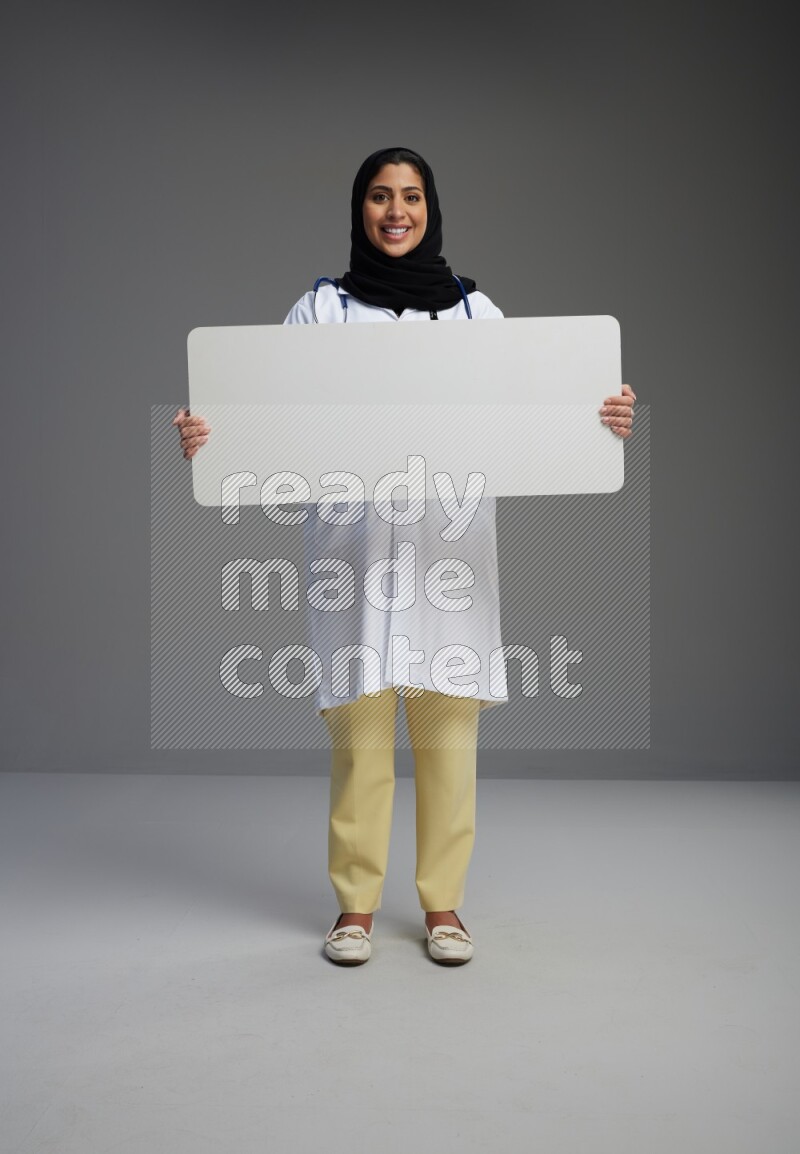 Saudi woman wearing lab coat with stethoscope standing holding board on Gray background