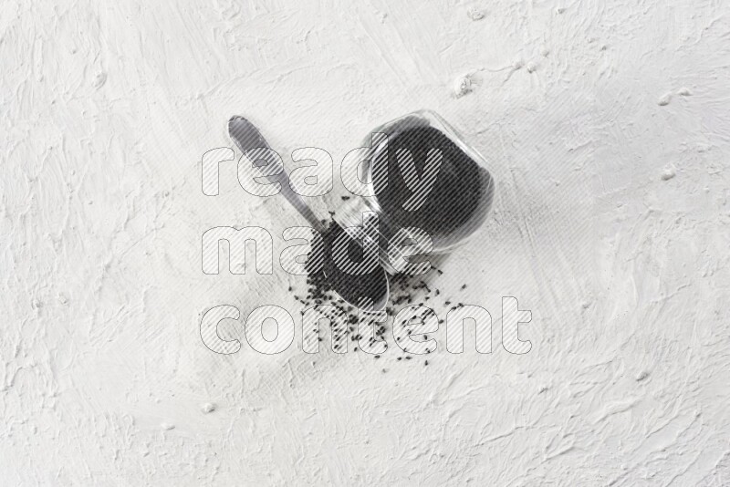 A glass spice jar and a metal spoon full of black seeds and the jar flipped and seeds spread on a textured white flooring