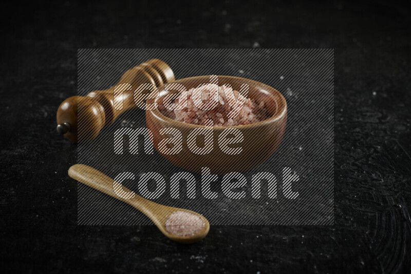 A wooden bowl and spoon filled with coarse pink himalayan salt and a wooden grinder beside them on black background