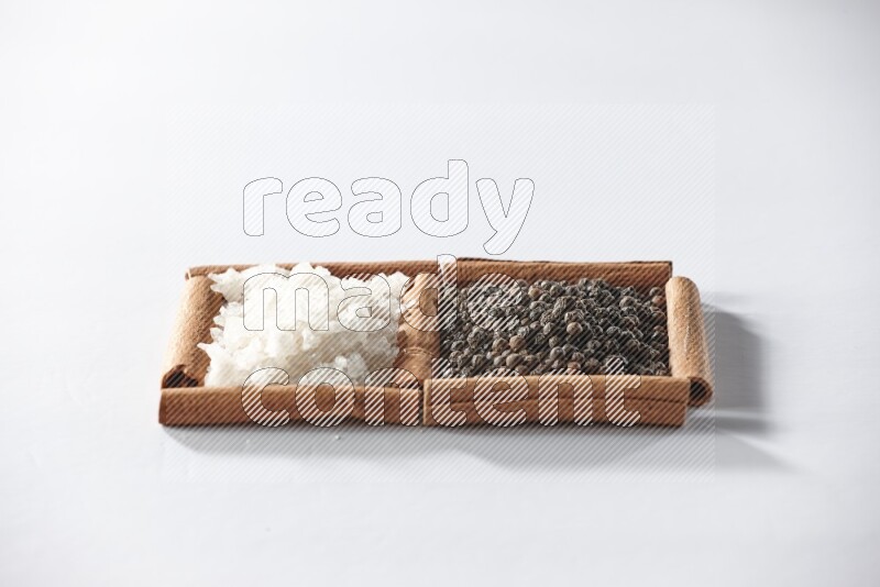 مربعان من أعواد القرفة ممتلئان بحبوب الفلفل الأسود والملح الأبيض علي خلفية بيضاء