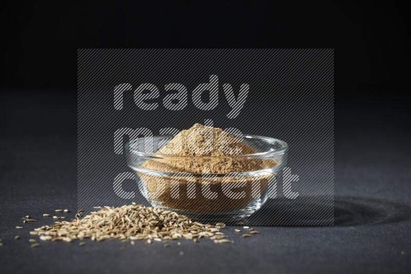 A glass bowl full of cumin powder with cumin seeds beside it on black flooring