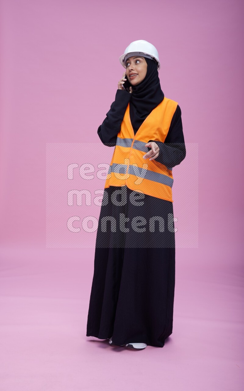 Saudi woman wearing Abaya with engineer vest and helmet standing talking on phone on pink background
