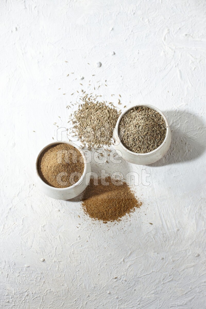 2 beige bowls full of cumin seeds and powder with spilled powder and seeds on textured white flooring