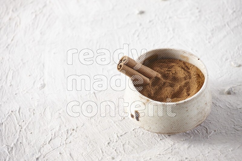 Ceramic beige bowl full of cinnamon powder with a cinnamon stick on a textured white background