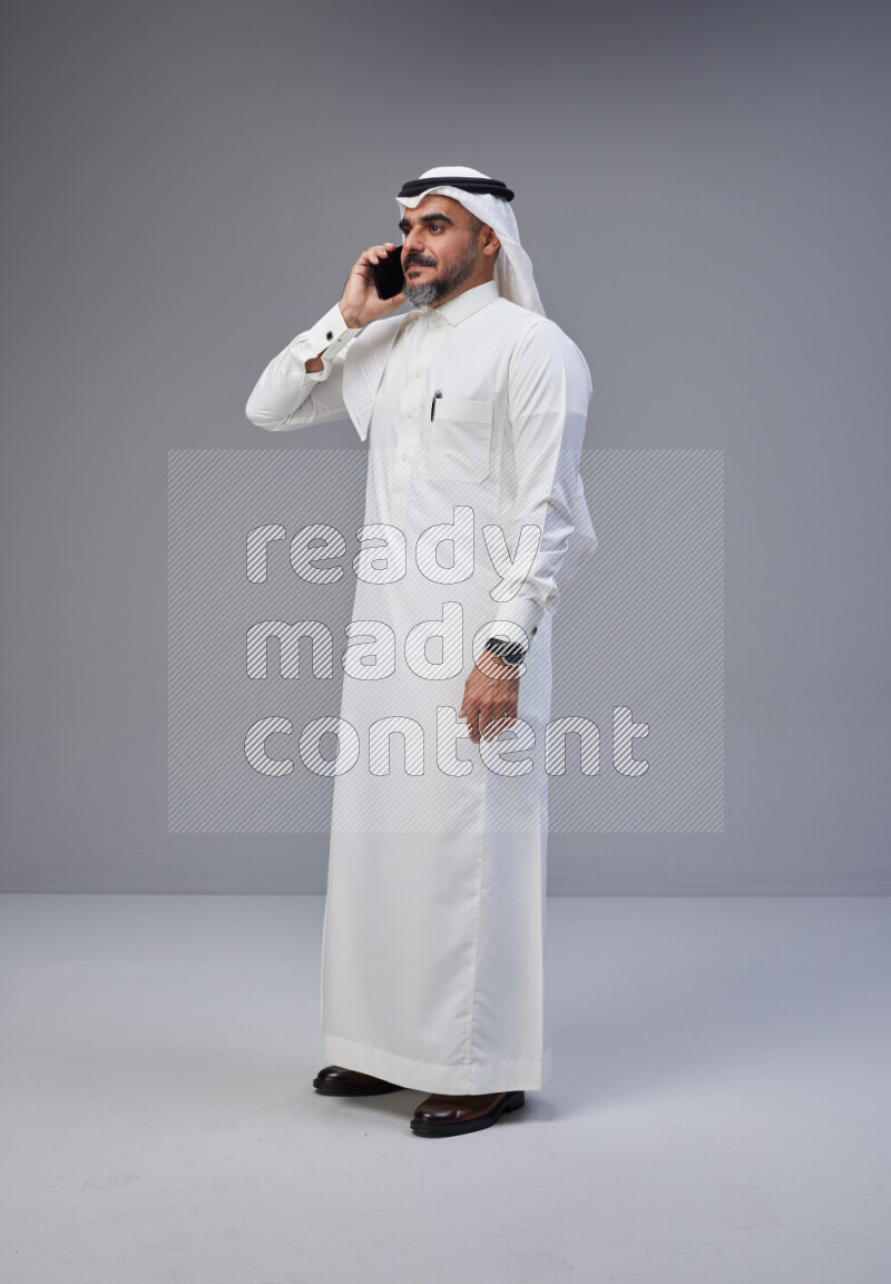Saudi man Wearing Thob and white Shomag standing talking on phone on Gray background