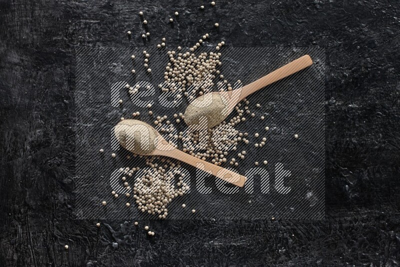 2 wooden spoons full of white pepper powder with white pepper beads on textured black flooring