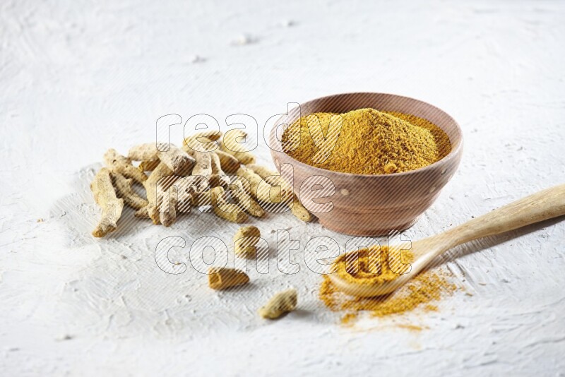 A wooden bowl and wooden spoon full of turmeric powder with dried turmeric fingers beside it on textured white flooring