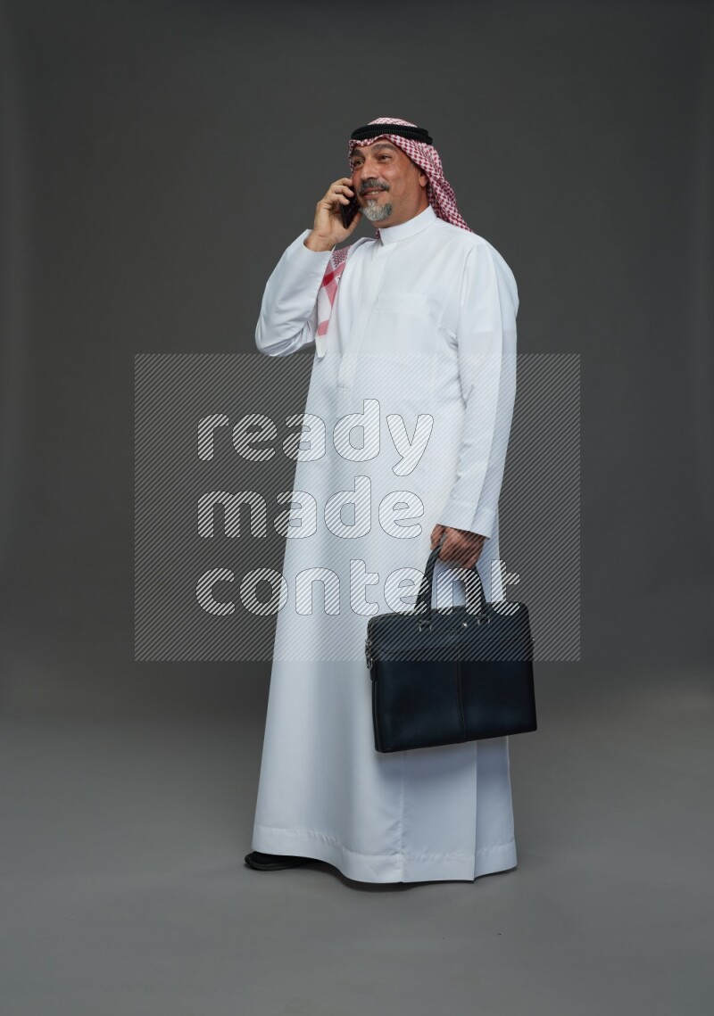 Saudi man with shomag Standing holding bag talking on phone on gray background