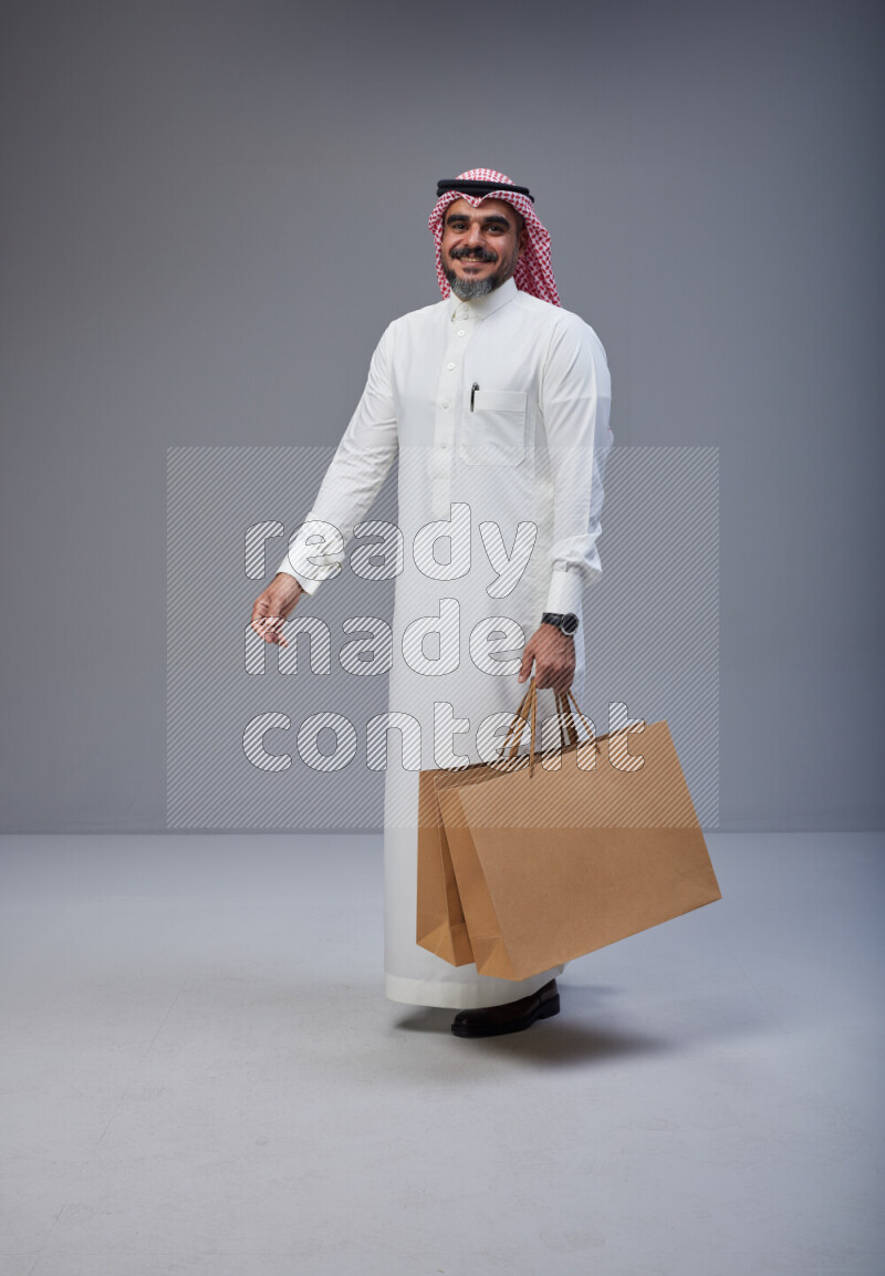 Saudi man Wearing Thob and red Shomag standing holding shopping bag on Gray background