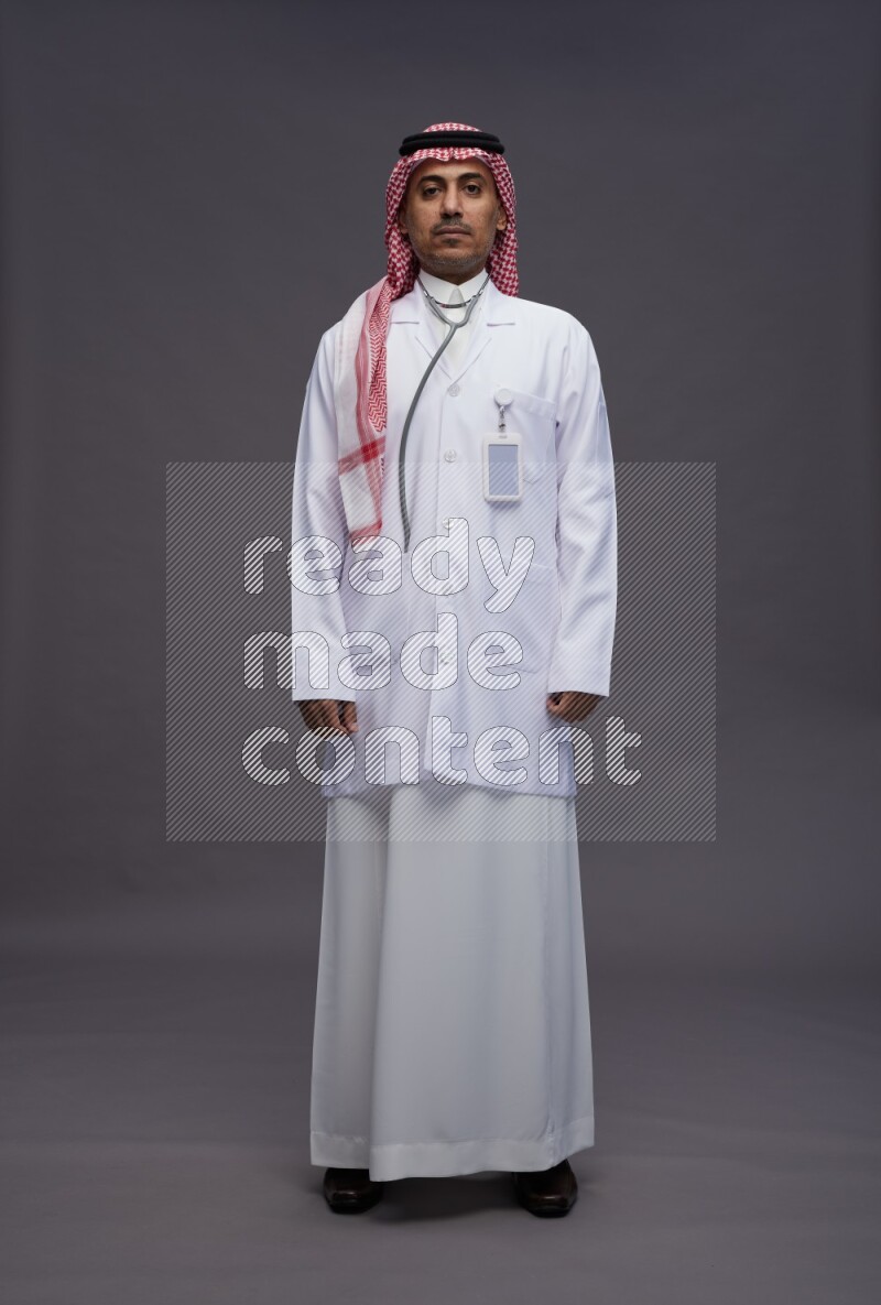 Saudi man wearing thob with lab coat and shomag with pocket employee badge with stethoscope standing interacting with the camera on gray background
