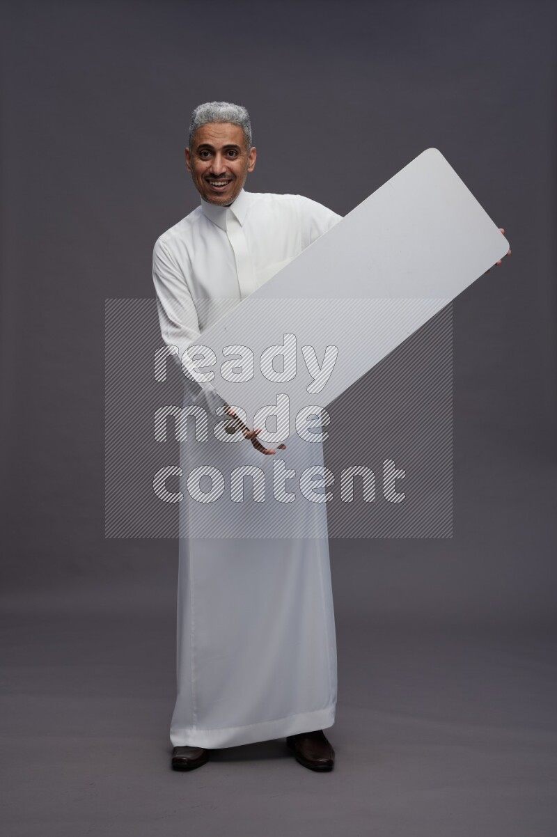 Saudi man wearing thob standing holding board on gray background