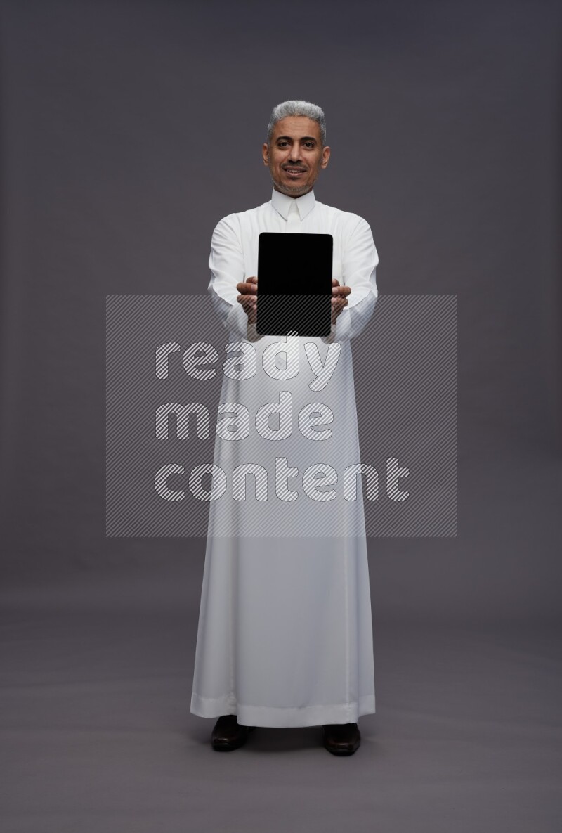 Saudi man wearing thob standing showing tablet to camera on gray background