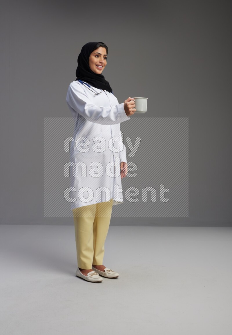 Saudi woman wearing lab coat with stethoscope standing holding mug on Gray background