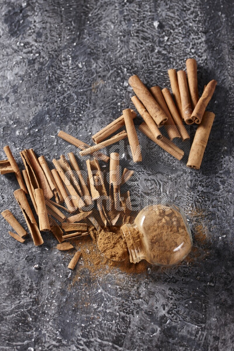 Herbal glass jar full cinnamon powder flipped and a metal spoon full of powder surrounded by cinnamon sticks on textured black background in different angles