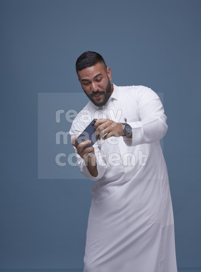 A man Playing a Game on his Smartphone  on Blue Background wearing Saudi Thob