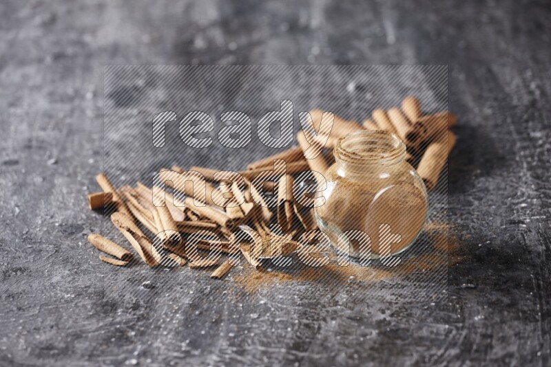 Herbal glass jar and a metal spoon full of cinnamon powder surrounded by cinnamon sticks on textured black background