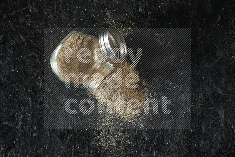 A flipped glass spice jar full of cardamom powder on textured black flooring