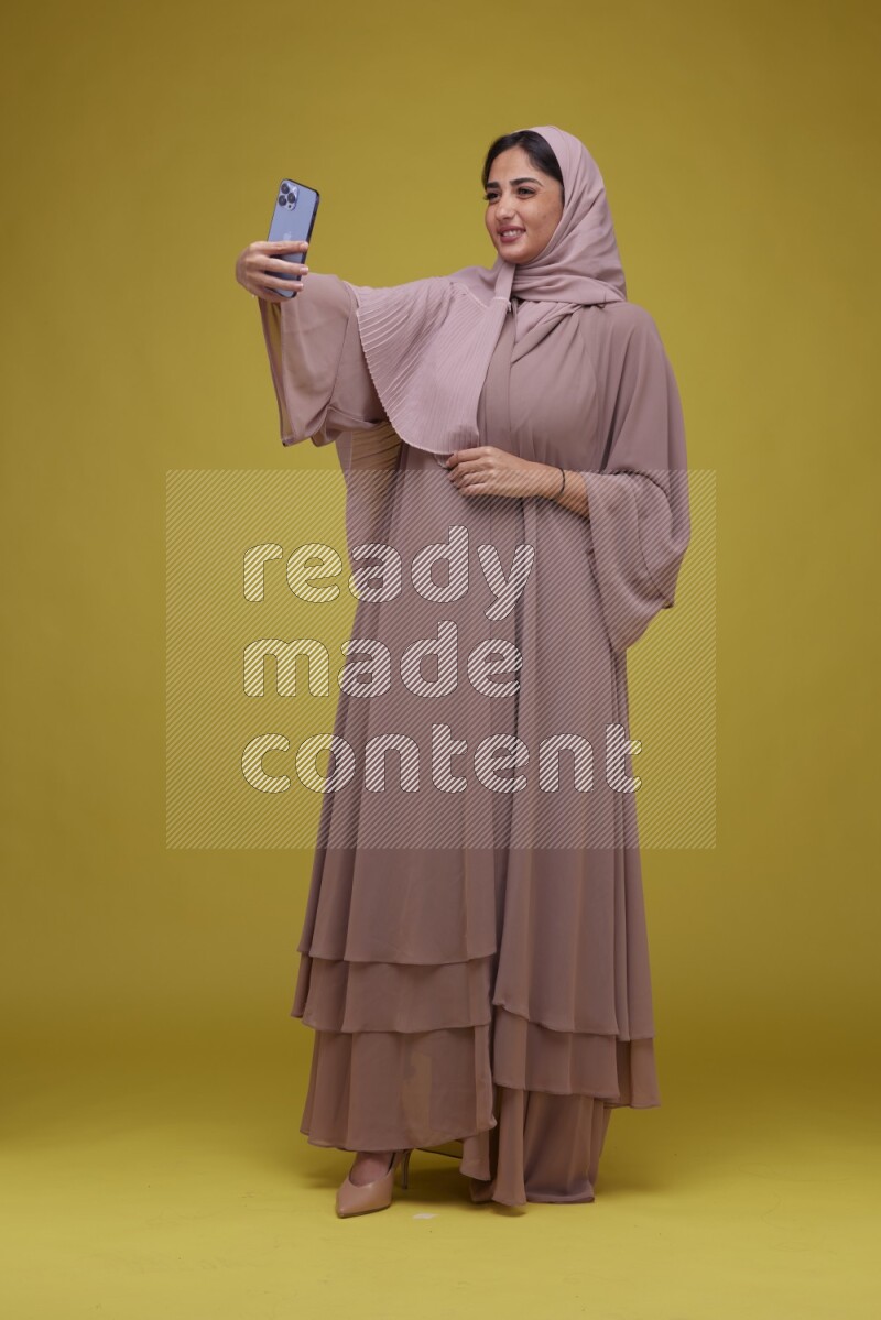 A woman taking a Selfie on a Yellow Background wearing Brown Abaya with Hijab
