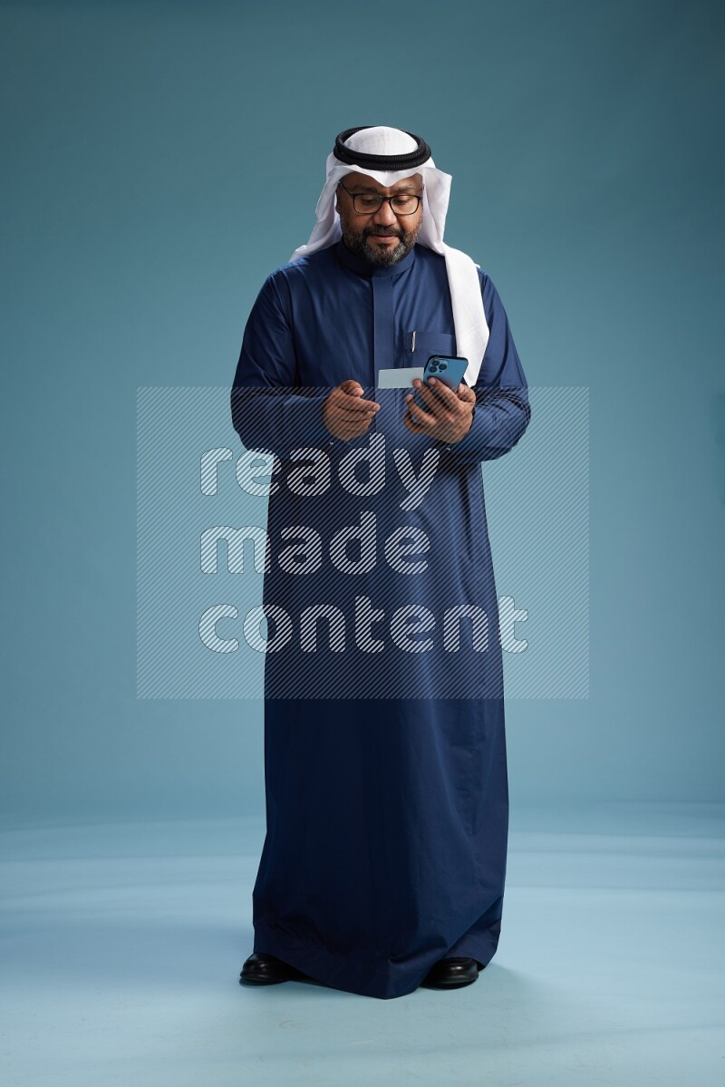 Saudi Man with shimag Standing holding ATM card while talking on phone on blue background