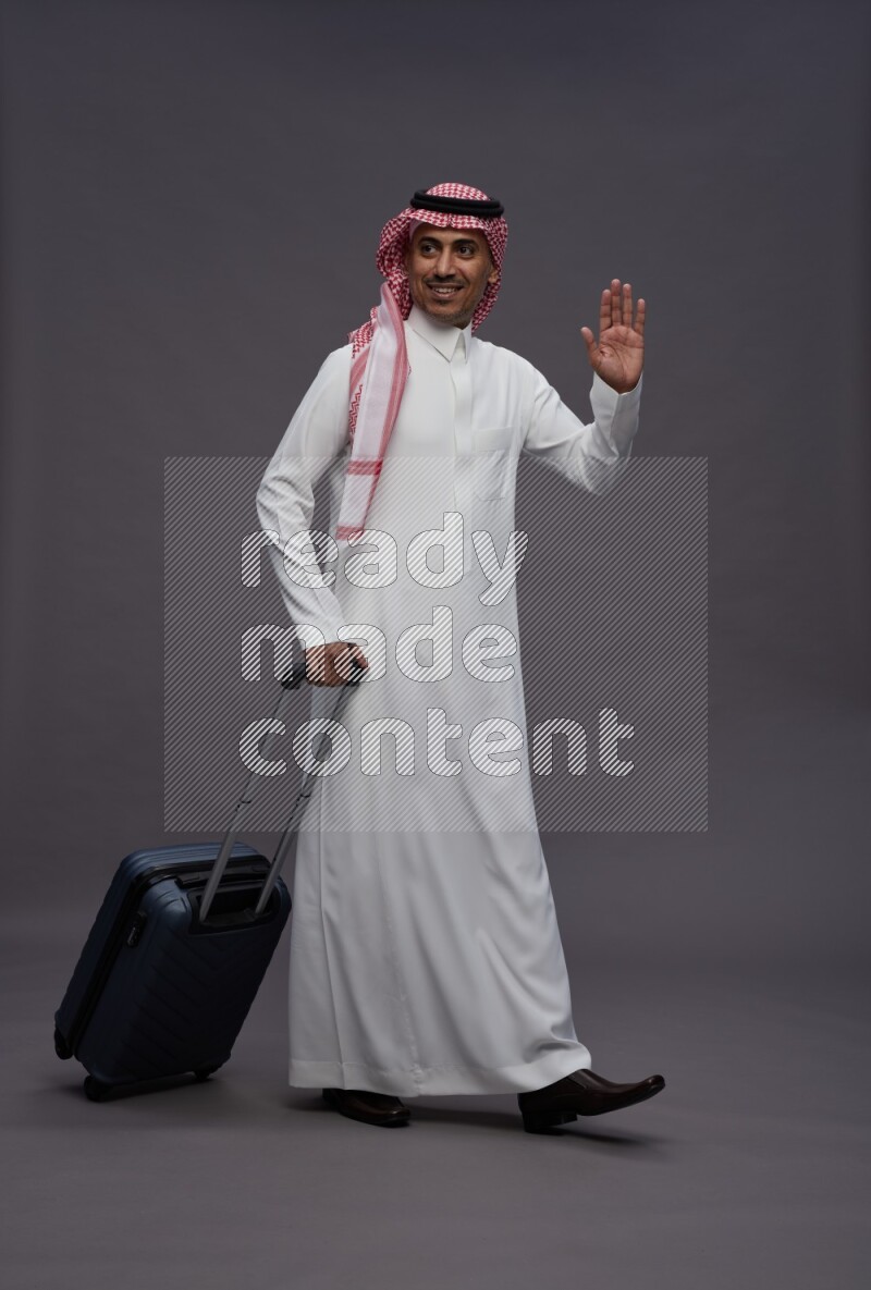 Saudi man wearing thob and shomag standing holding bag on gray background