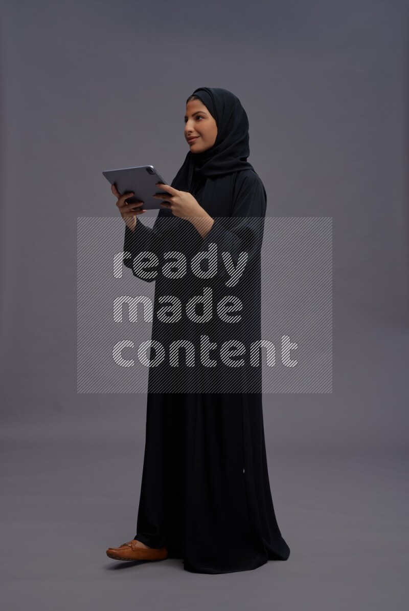 Saudi woman wearing Abaya standing working on tablet on gray background