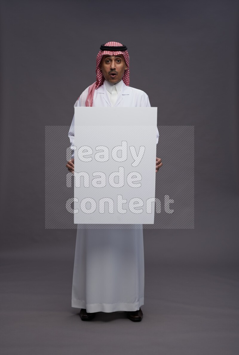 Saudi man wearing thob with lab coat and shomag with pocket employee badge standing holding board on gray background