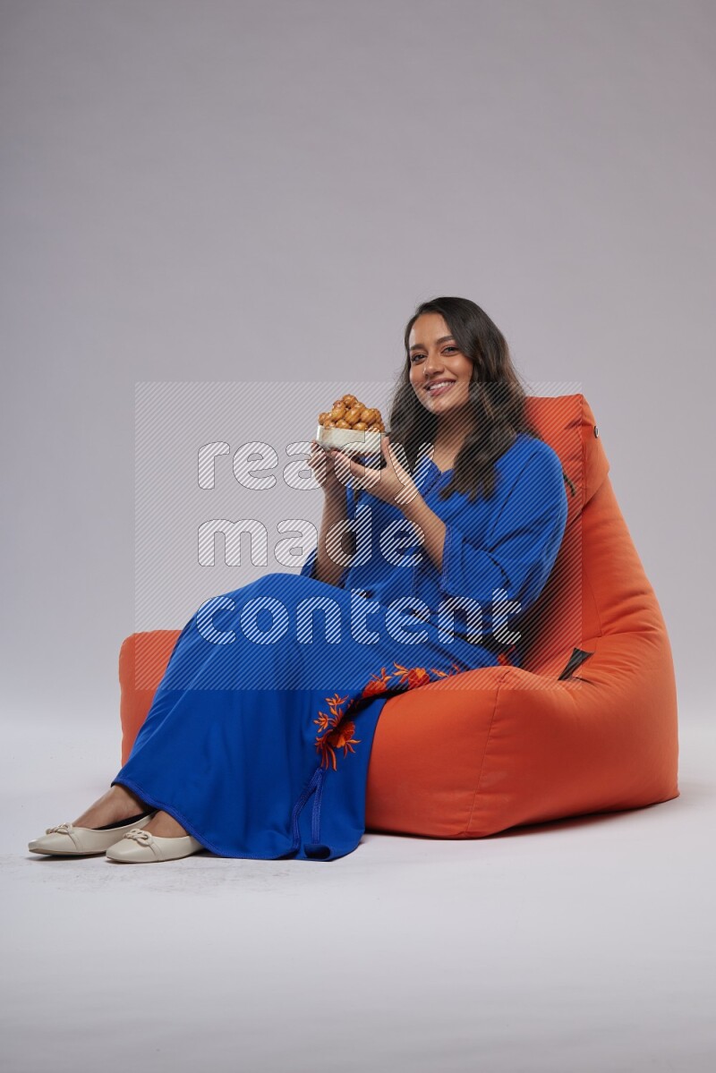 A Woman sitting on an orange beanbag wearing Jalabeya holding a plate of luqaimat