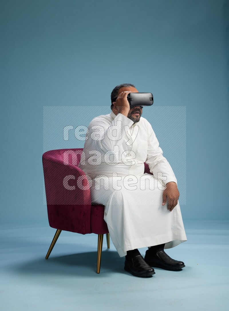 Saudi Man without shimag sitting on chair  gaming with VR on blue background