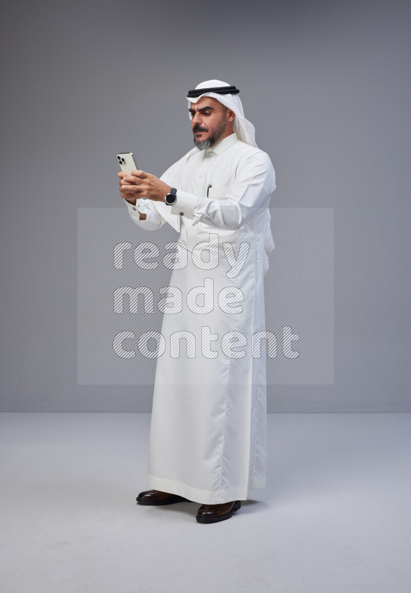 Saudi man Wearing Thob and white Shomag standing texting on phone on Gray background