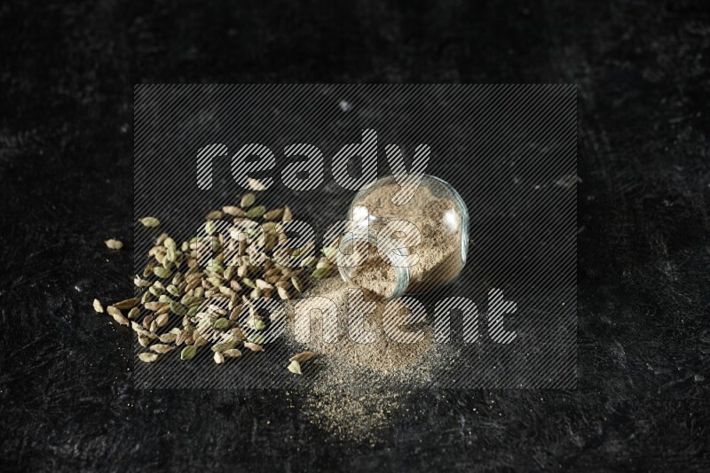 وعاء زجاجي للتوابل مقلوب وممتلئ ببودرة الهيل والبودرة تناثرت منه مع حبوب الهيل المتناثرة على خلفية سوداء