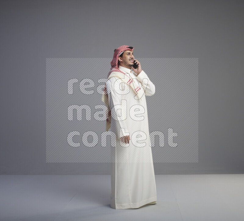 A Saudi man standing wearing thob and red shomag talking on phone on gray background