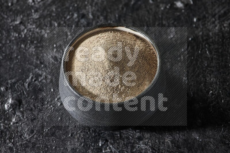 Black pottery bowl full of white pepper powder on textured black flooring