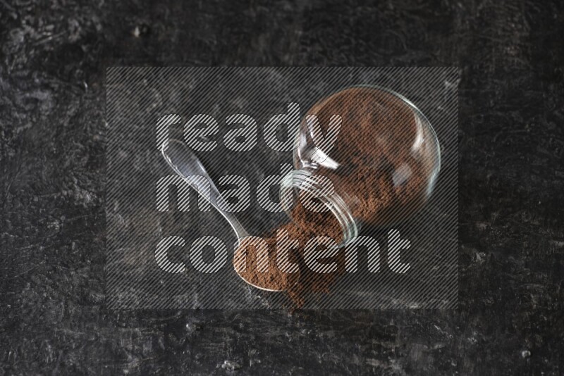 A flipped glass spice jar and a metal spoon full of cloves powder on textured black flooring