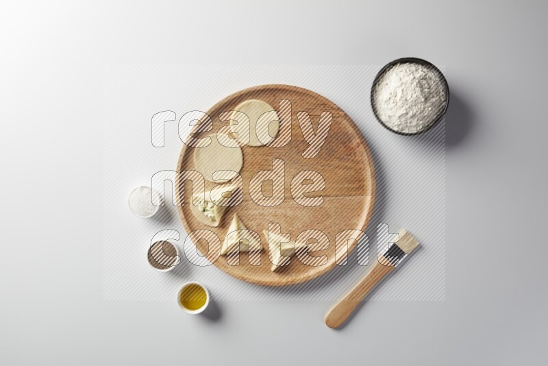 two closed sambosas and one open sambosa filled with cheese while flour, salt, black pepper and oil with oil brush aside in a wooden dish on a white background