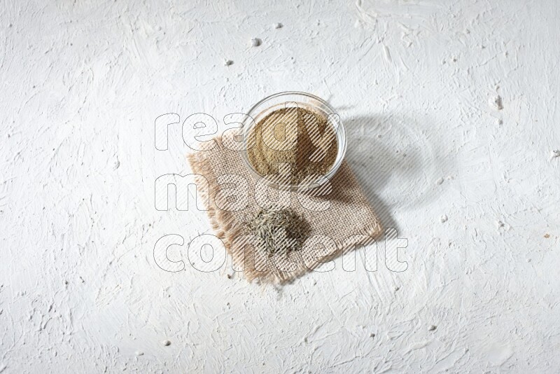 A glass bowl full of cumin powder with some of cumin seeds on burlap piece on a textured white flooring