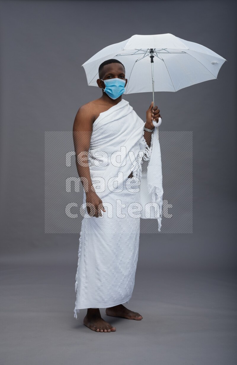 A man wearing Ehram with face mask Standing holding umbrella on gray background
