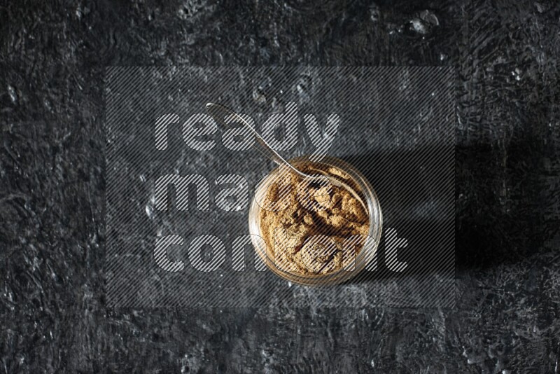 A glass jar and a metal spoon full of allspice powder on a textured black flooring