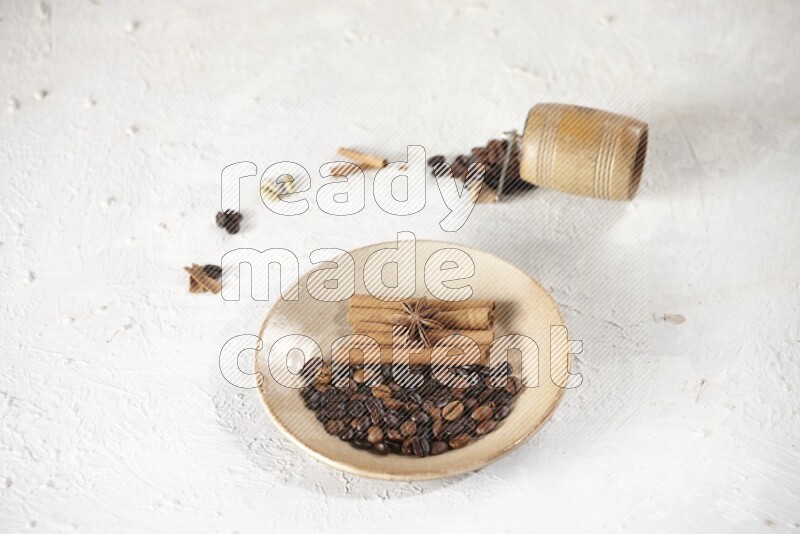 Beige plate full of coffee beans, cinnamon sticks and star anise with a coffee grinder, coffee beans, cinnamon pieces and cardamom next of it on white background