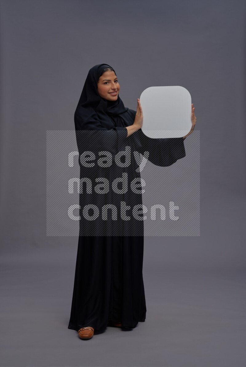 Saudi woman wearing Abaya standing holding social media sign on gray background