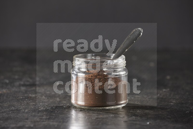 A glass jar full of cloves powder with a metal spoon on a textured black flooring