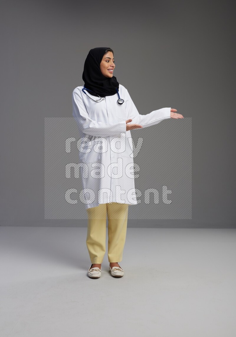 Saudi woman wearing lab coat with stethoscope standing interacting with the camera on Gray background