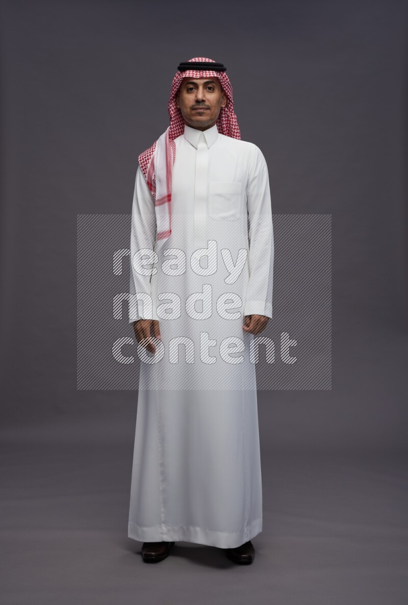 Saudi man wearing thob and shomag standing interacting with the camera on gray background