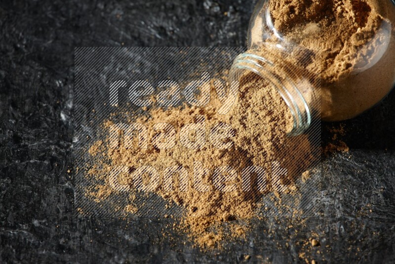 A flipped glass spice jar full of allspice powder and powder spilled out of it on a textured black flooring